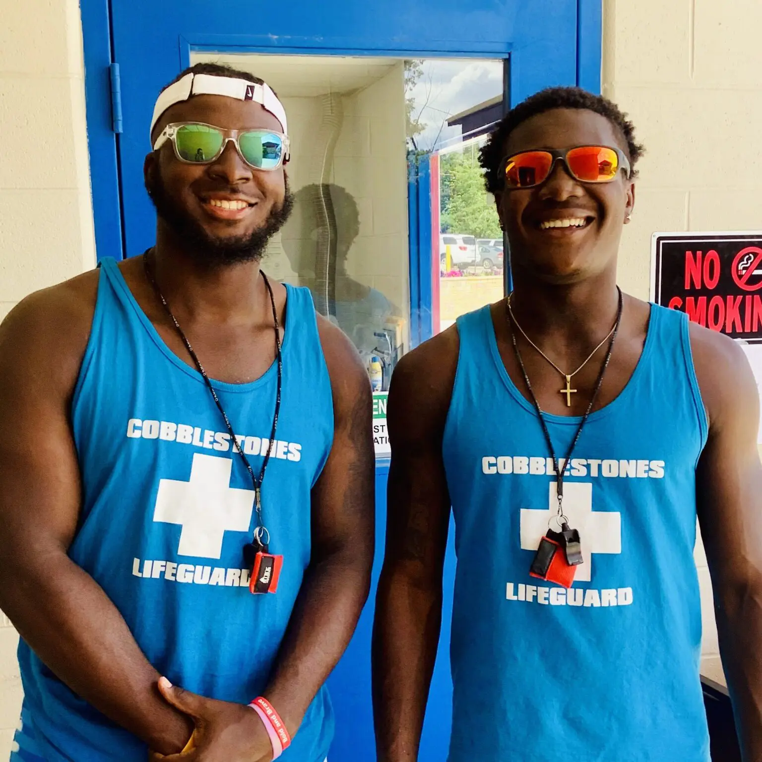 Two lifeguards smiling, wearing matching blue tank tops and sunglasses.